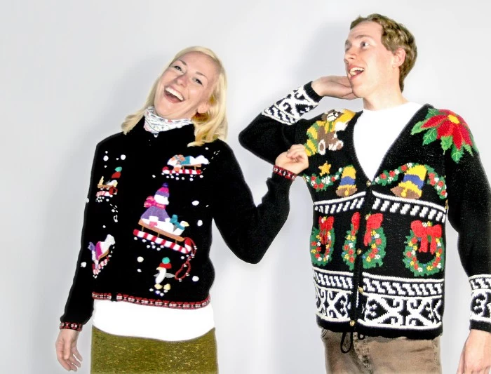 straw blonde woman, laughing and wearing a black festive jumper, with colorful sleds and snowmen, near a laughing man, in a black cardigan, decorated with bells and christmas wreaths