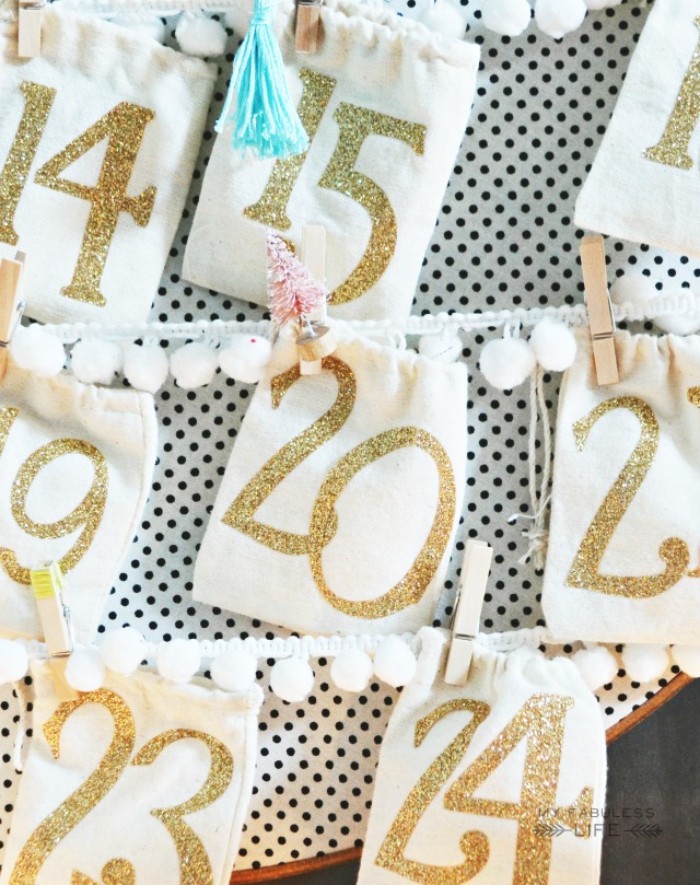pale cream pouches, decorated with numbers, written in gold glitter, seen in close up, christmas advent calendar, each pouch is attached to white lace ribbon, with a miniature clothes peg