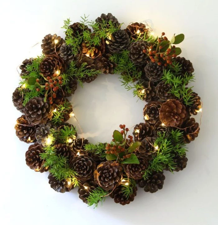 glowing string lights, decorating a christmas wreath, made from pinecones, and little green leaves, with small berries, on a white surface