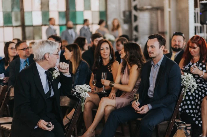 guests at a wedding, or another formal event, sitting on chairs, some with champagne flutes in their hands, dressed in black tie optional attire, black and dark navy suits, with white and light colored shirts, smart occassion dresses