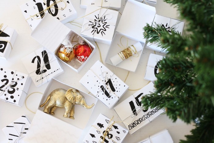 white boxes with numbers, written on them in black, some open and some closed, the open ones contain a metallic, gold elephant figurine, christmas tree ornaments, and other items, advent calendar ideas, fake christmas tree branches nearby