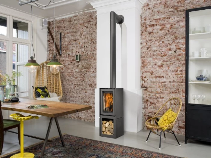 7 tips to make your home cosy, modern metal fireplace in black, with a firewood compartment, in a room with a brick wall, smooth light grey floor, wooden table and a black cupboard, rattan swing and chair