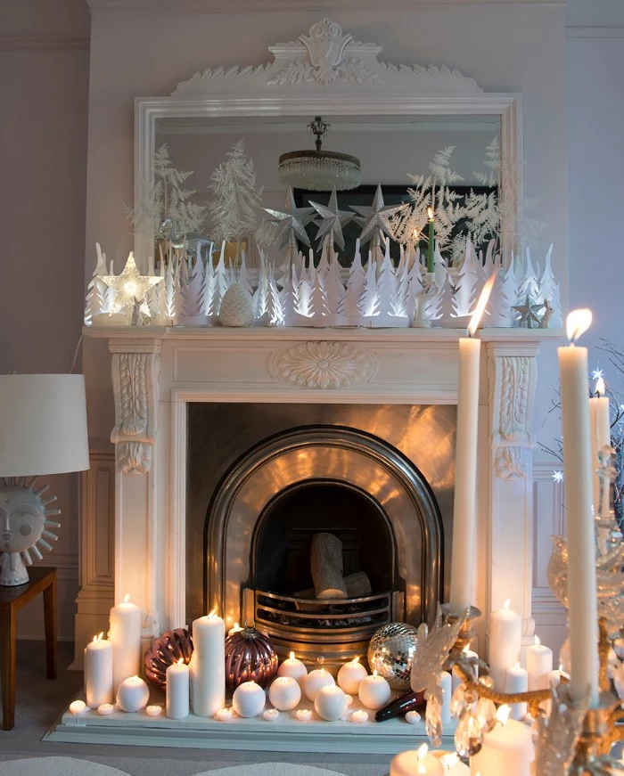 lots of burning white candles, in different shapes and sizes, near a vintage-style fireplace, in white and silver, decorated with multiple white, illuminated christmas tree figurines, christmas fireplace