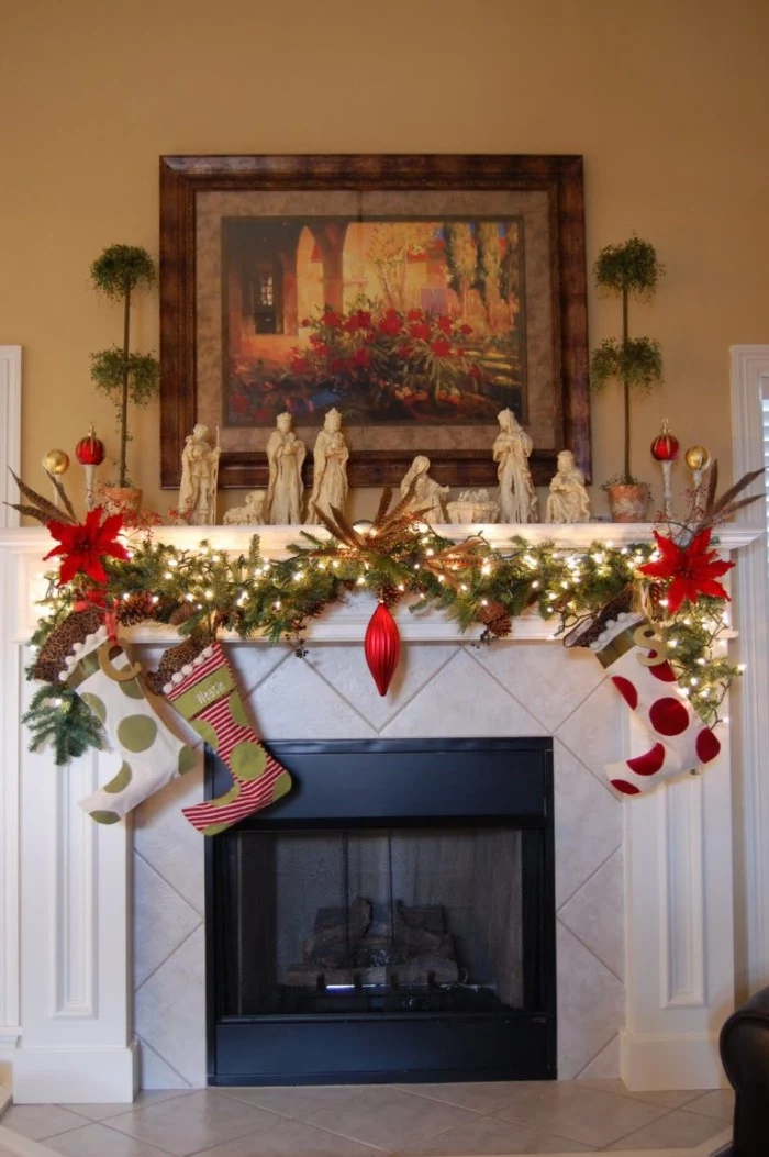poinsettia flowers and a red ornament, on a pine garland, decorated with glowing fairy lights, and three stockings, mantel decor, with a nativity scene