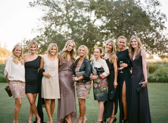 group photo of nine smiling blonde women, dressed in cocktail attire, maxi and midi dresses in black, white and ash pink, and featuring floral patterns