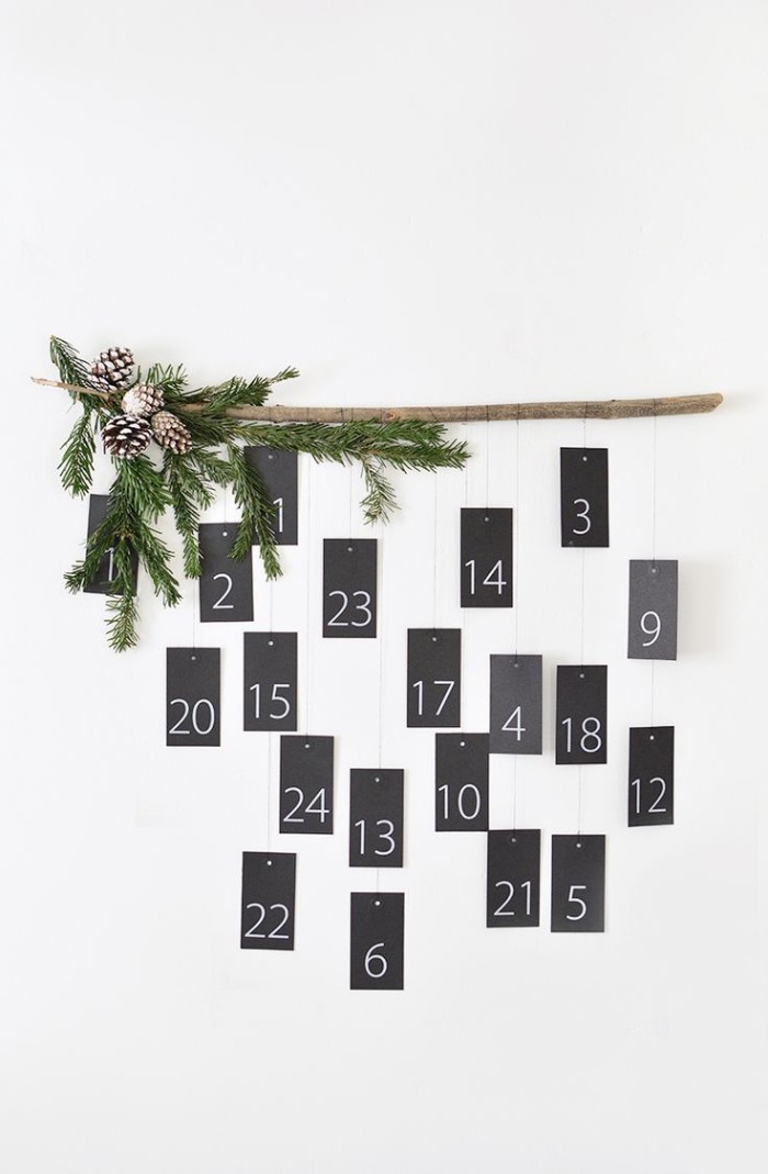 black cards hanging on pieces of string, from a wooden pole, decorated with pine cones, and fir branches, each card has a white number, from 1 to 24, christmas countdown calendar