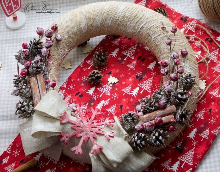 holiday wreaths, close up of a wreath, wrapped in beige string, and decorated with pinecones, red faux berries, cinammon sticks and a pale beige bow, with a large red snowflake ornament, all dusted in faux snow