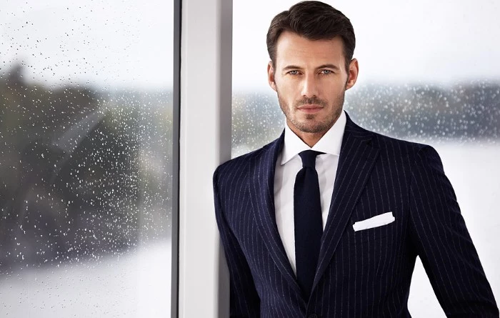 navy blue suit, with pinstripe pattern, worn by a brunette man, with stubble on the lower part of his face, cocktail attire for men, window with rain drops in the background