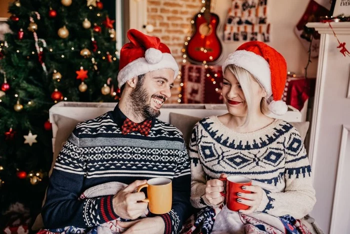 ugly christmas sweater ideas, couple wearing patterned winter jumpers, and santa hats, smiling while holding mugs