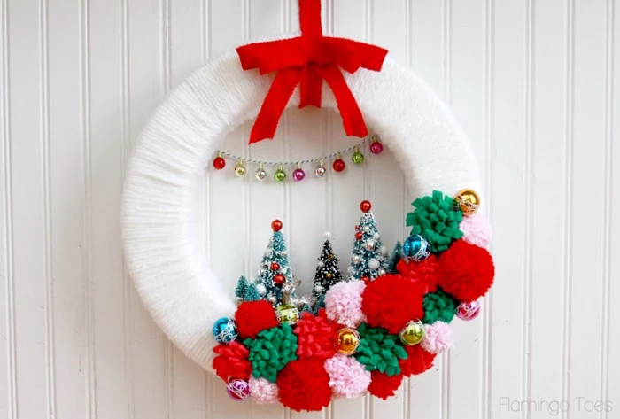 white yarn wrapped around a broad hoop, decorated with red, green and pale pink pompoms, three christmas tree figurines, miniature baubles on a silver chain, and a red bow, diy christmas wreath