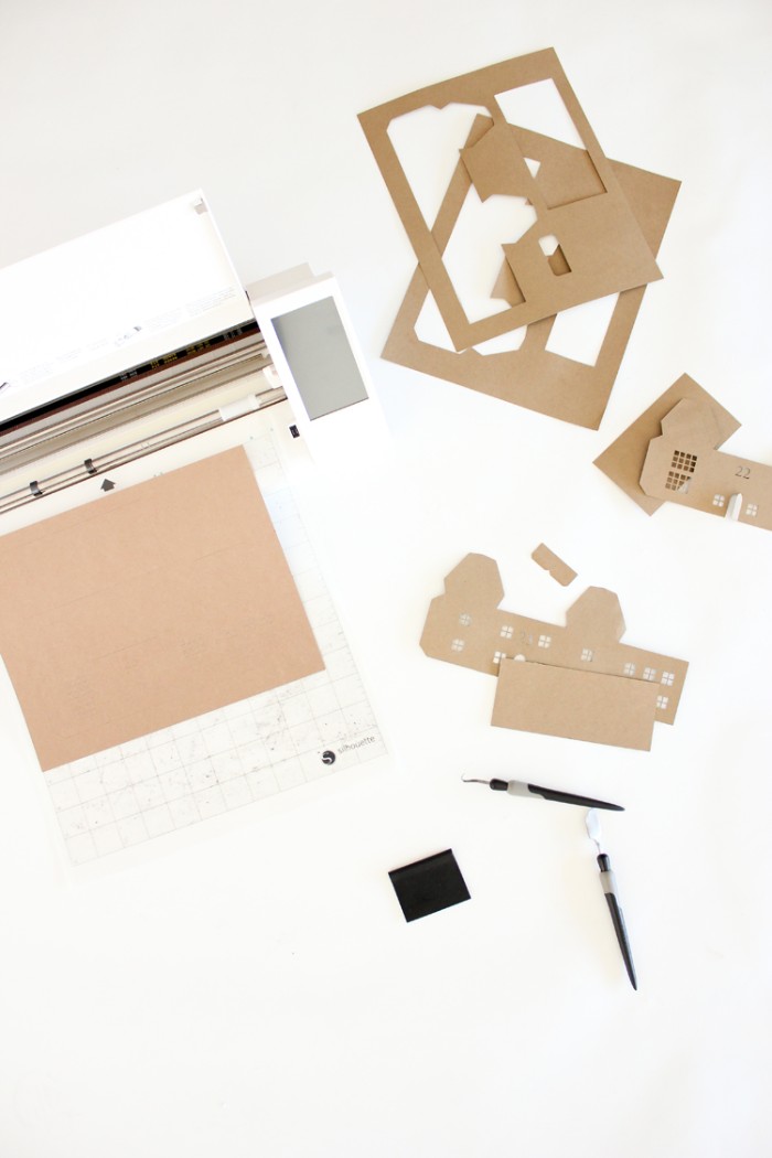 paper trimmer and a piece of beige card, on a white surface, two small utility knives, and several beige card cutouts, making an adult advent calendar 