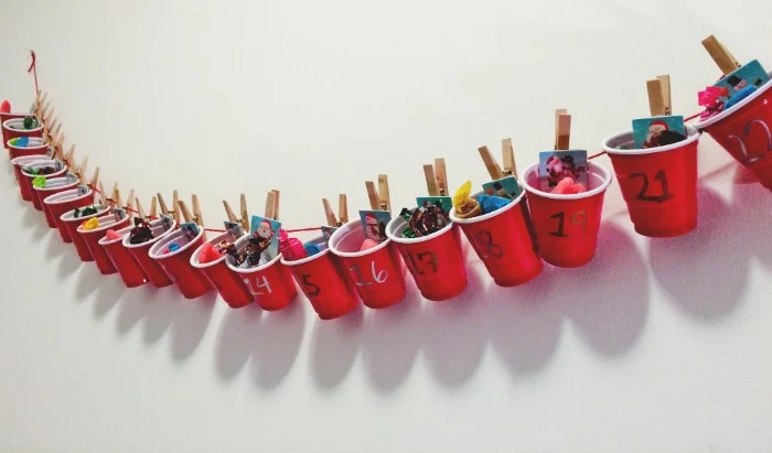 plastic cups in red and white, filled with candy and small gifts, attached with wooden clothes pegs, to a red piece of string, hanging on a white wall