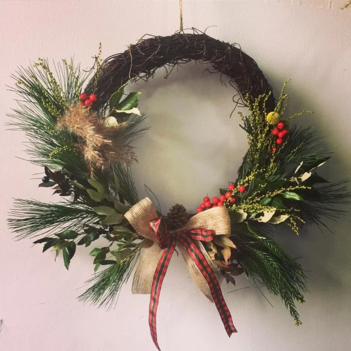 pine leaves and sprigs from other green, and dried plants, decorating a rustic-style wreath, made from thin, dried and twisted branches, and adorned with bows, and a pinecone