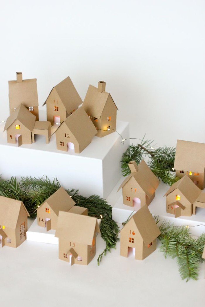 pine branches and lit string lights, placed on a white survace, among thirtheen decorations, shaped little houses, made from beige card, adult advent calendar suggestion