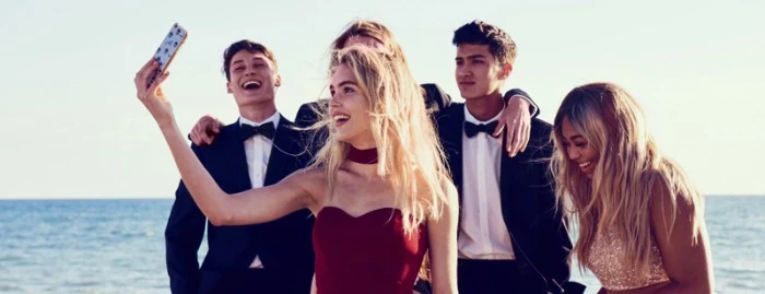 group of laughing teenagers, standing by the sea, blonde young woman, dressed in a dark red gown, holding her phone up for a selfie