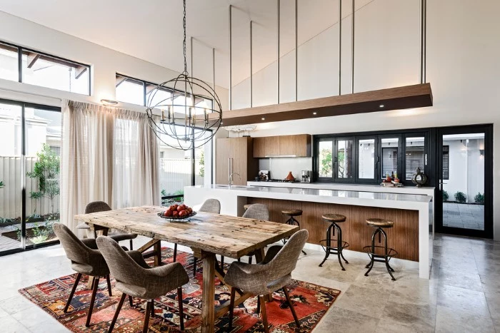 open plan dining area and kitchen, featuring a rustic wooden table, several mink brown chairs, a kilim rug, and large double windows, kitchen island with bar chairs