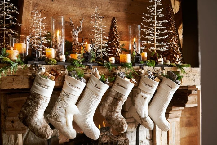 delicate ornaments shaped like thin, white christmas trees, burning candles and a leafy green garland, on a rustic wooden fireplace, mantel decor, six white and beige stockings 