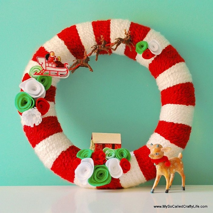 red and white, striped christmas wreath, decorated with small figurines, santa's reindeer sleigh, a little house, felt flowers in white, red and light green, holiday wreaths, a small deer toy, with a red scarf
