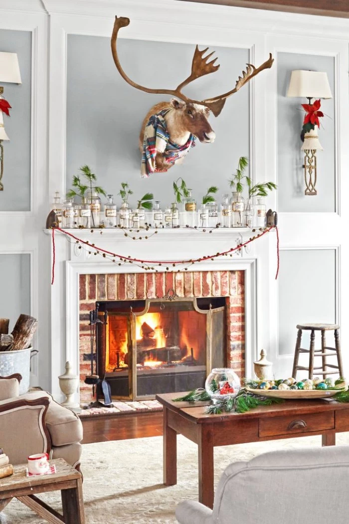 moose's head stuffed, and decorated with a winter scarf, mounted on a grey and white wall, above a fireplace, decorated with multiple jars, containing pine branches, holiday images, burning fire and festive ornaments