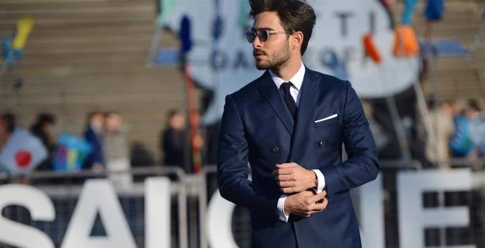smart navy suit, worn over a white shirt, and a black tie, by a young man with sunglasses, a moustache and stubble on his chin, cocktail attire ideas for guys