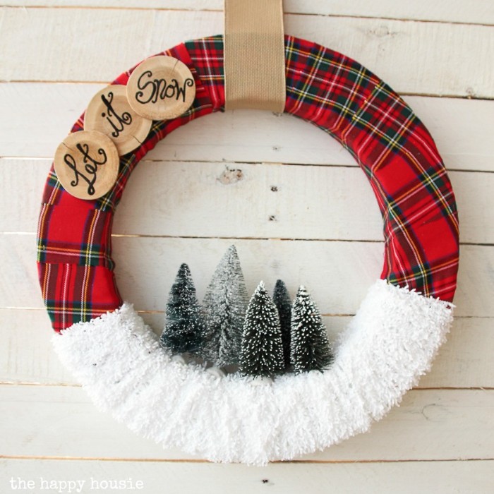let it snow, written in black, on three round wooden tags, stuck onto a wreath, covered in red tartan fabric, and white gauze, christmas wreath images, five snowy christmas tree figurines in the middle