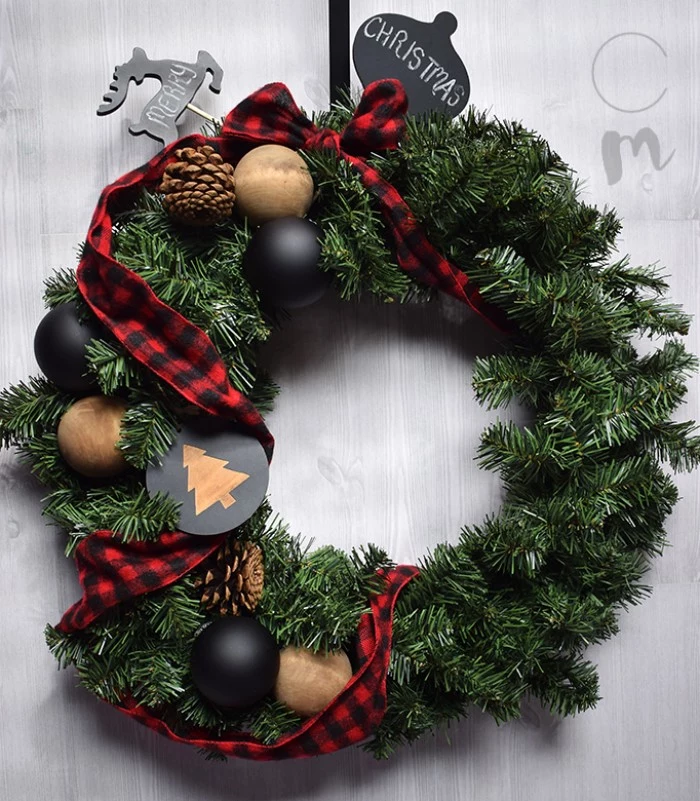 fake fir diy christmas wreath, decorated with black and beige christmas tree baubles, pinecones and a red and black, checkered ribbon tied into a bow, small black shapes, with the words merry and christmas, written in white chalk