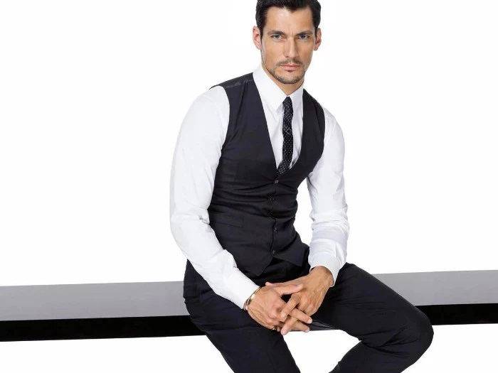 cocktail attire for men, brunette man with short beard and mustache, sitting on a black surface, dressed in black trousers, a smart black vest, white shirt and a dark patterned tie