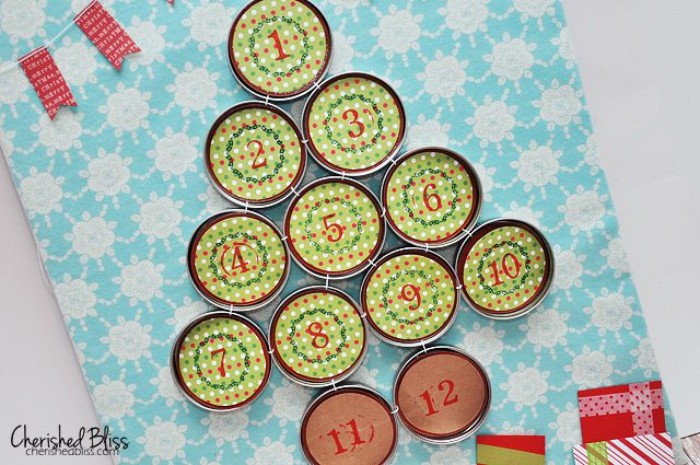 numbered mason jar lids, decorated with patterned, light green and red paper, organized in the shape of a christmas tree, fun advent calendars, on a light blue and white surface, with a floral pattern