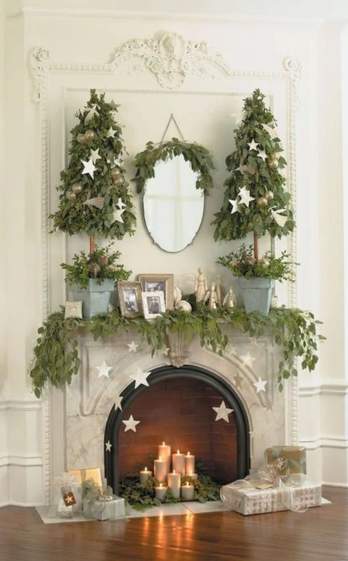 star-shaped ornaments in silver, suspended from the mantelpiece, of a classic fireplace, covered in green garlands, made of leaves, fireplace mantel decor, burning candles and two small christmas trees