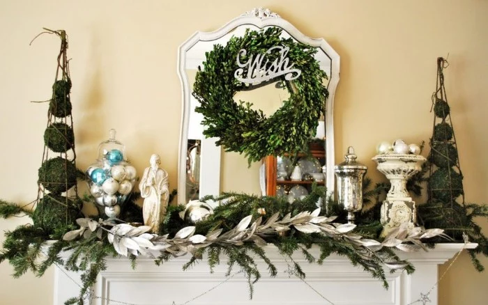 wish written in silver, on an ornament, decorating a simple green wreath, hung over a mirror, above a white mantelpiece, holiday images, pine sprigs and a decorative branch with silver leaves