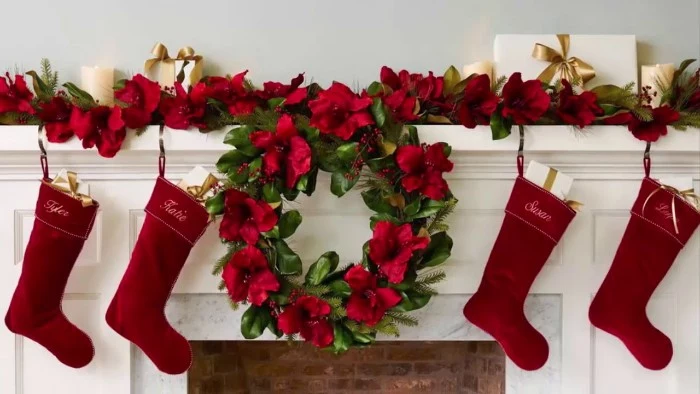 burgundy velvet stockings, each inscribed with a name, hanging on a white mantelpiece, near a green wreath with faux red flowers, christmas mantel ideas, similar red and green garland in the background