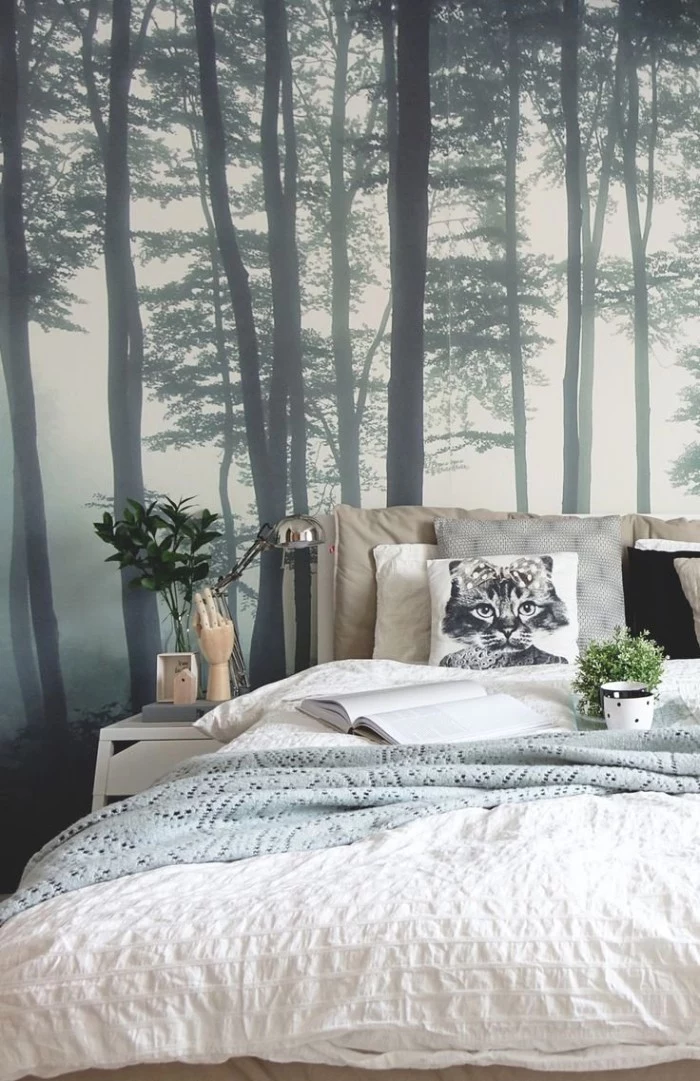 misty forest scene, in dark grey and white, on a wallpaper behind a bed, covered in white, grey and beige cushions, duvet and blanket