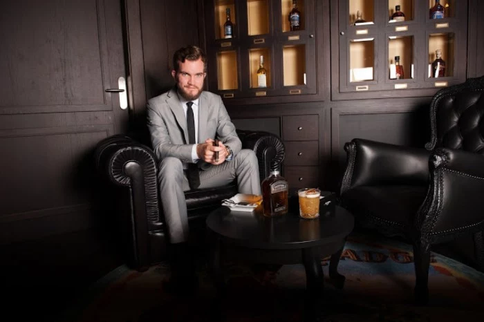 smiling man in a light grey suit, with a white shirt, and a dark tie, sitting on a black leather armchair, near a small wooden table, with drinks and a snack, cocktail attire examples for men