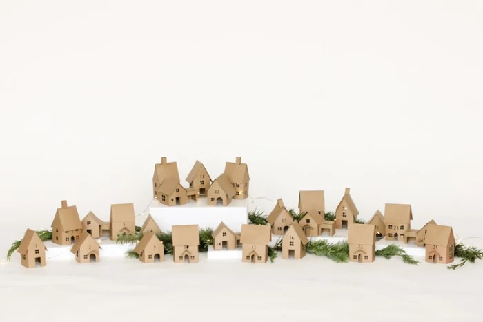 multiple small houses, made from beige card, placed on a white surface, decorated with lit string lights, and pine branches, adult advent calendar diy, decorative christmas village