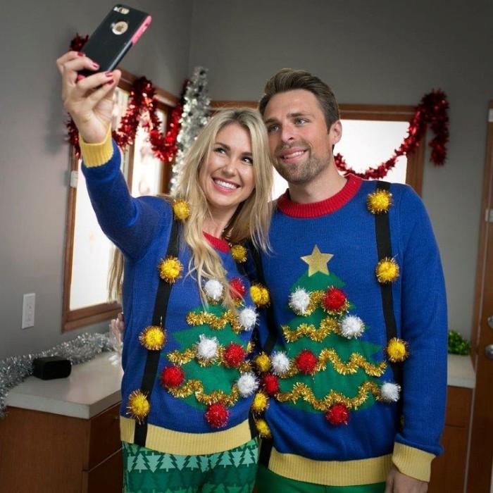 identical blue jumpers, with christmas trees, decorated with gold garlands, and white and red pom poms, worn by a smiling couple, posing for a selfie