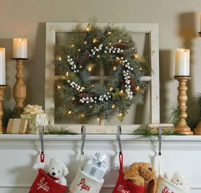 wooden candlesticks holding three lit candles, near a christmas wreath, decorated with small glowing fairy lights, and white faux berries, christmas mantel ideas