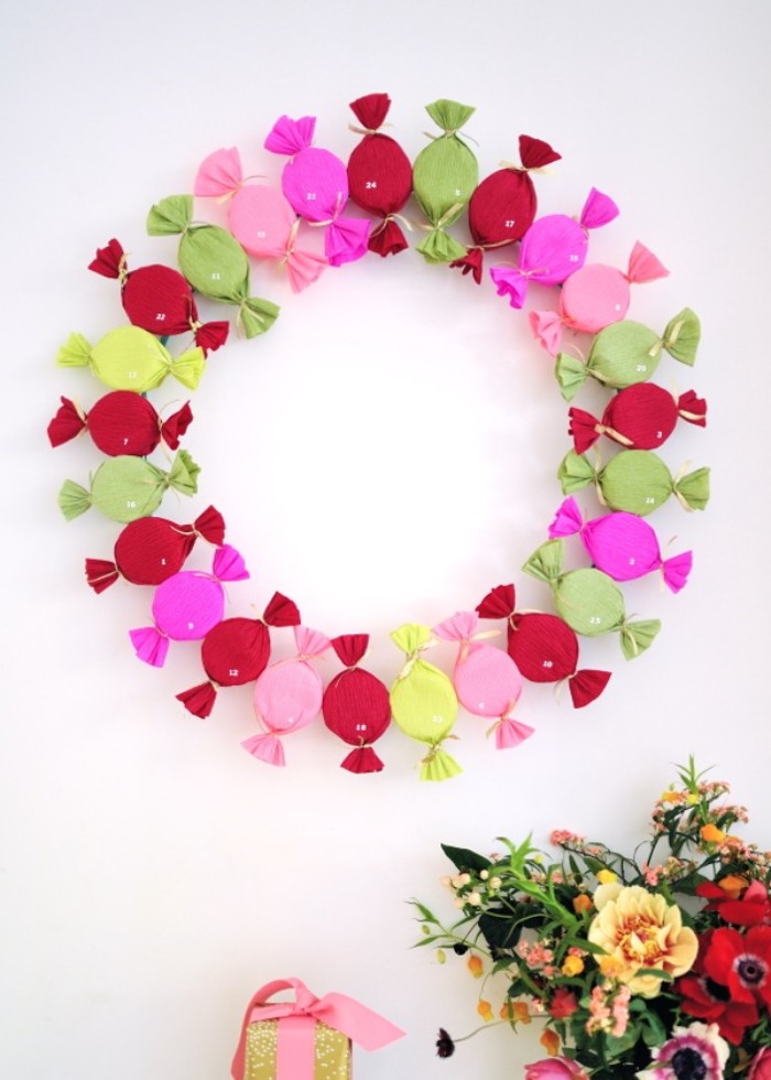 decorative festive freath/ advent calendar, made from small, mutlicolored candy-shaped presents, mounted on a white wall, near a flower bouquet