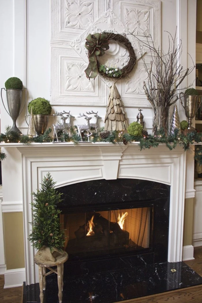 minimalistic diy fireplace mantel decoration, featuring a handmade, wooden christmas tree figurine, a vase with dried branches, four silver reindeer statuettes, pine leaves and pine cones