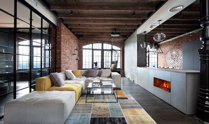 brick walls, dark wooden floor, large white and yellow couch with white and yellow cushions, geometrical blue, grey, yellow and black carpet, fireplace, room ideas