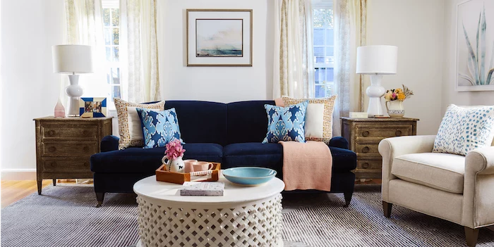 white walls, wooden floor with grey carpet, blue velvet sofa with printed blue and beige throw pillows, round white coffee table, interior design ideas