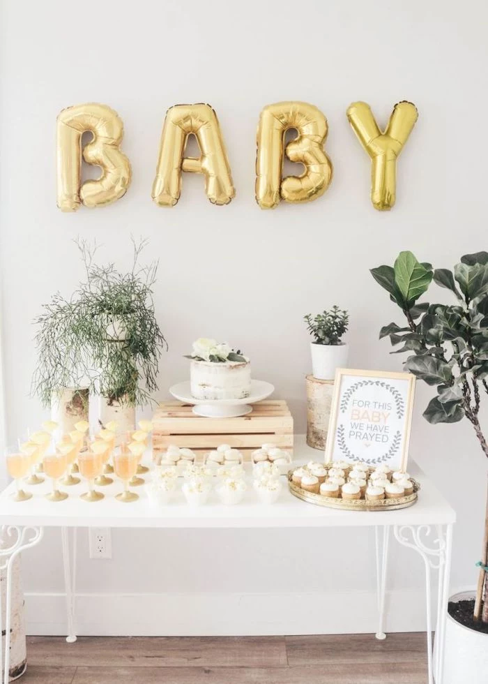 baby gold balloons, cake and sweets on the table, white backdrop, baby shower themes for boys, flowers in pots
