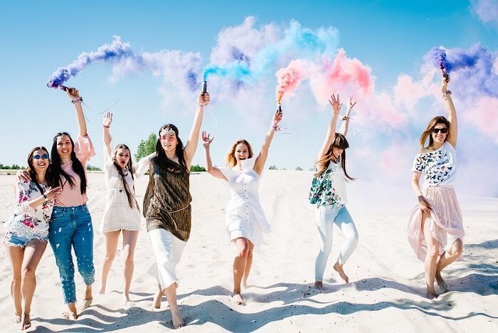 group of women on the beach, colour bombs in the air, feet in the sand, bachelorette party ideas