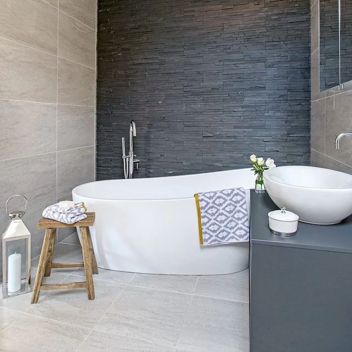grey stone tiled wall, bathroom shower ideas, white bathtub and sink, grey cabinet, wooden stool