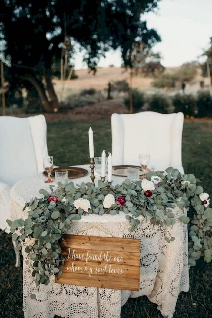 table with green and white roses flower arrangement, candles in candlesticks, white armchairs, diy wedding decorations