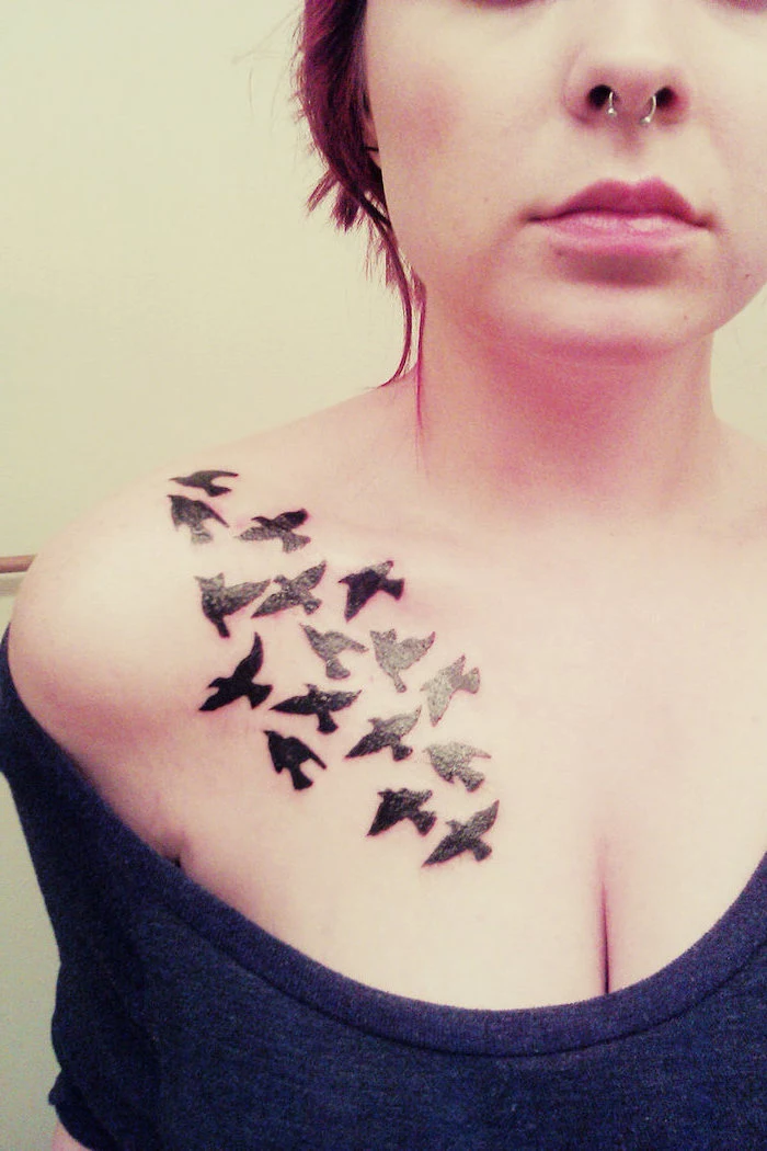grey top, white background, birds flying on shoulder, tribal chest tattoos, nose earring, female neck and chest tattoos
