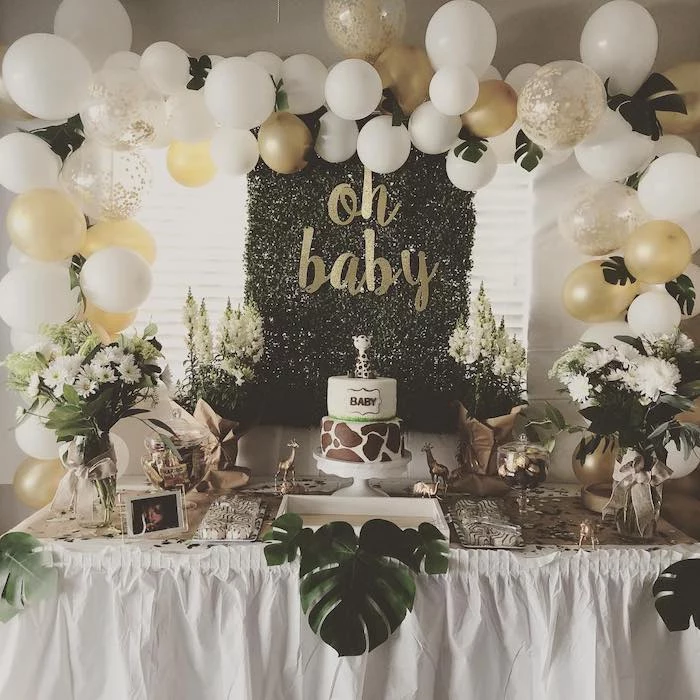 white and gold balloons arch, cake and sweets on the table, baby shower theme ideas, flower bouquets in vases
