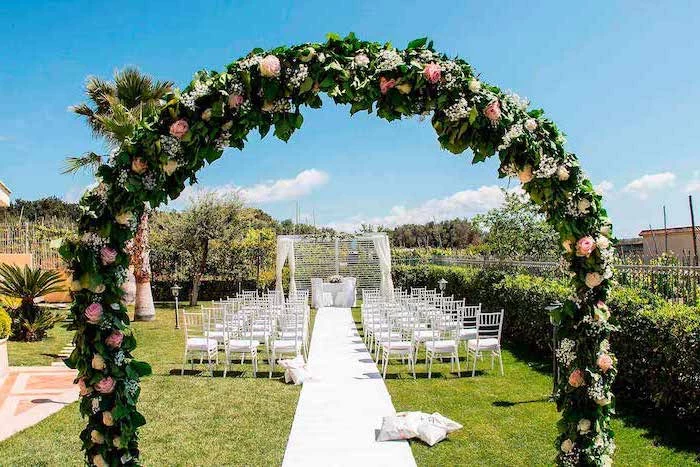 pink and yellow roses arch, white chairs and tulle next to the aisle, palm trees, wedding decoration ideas