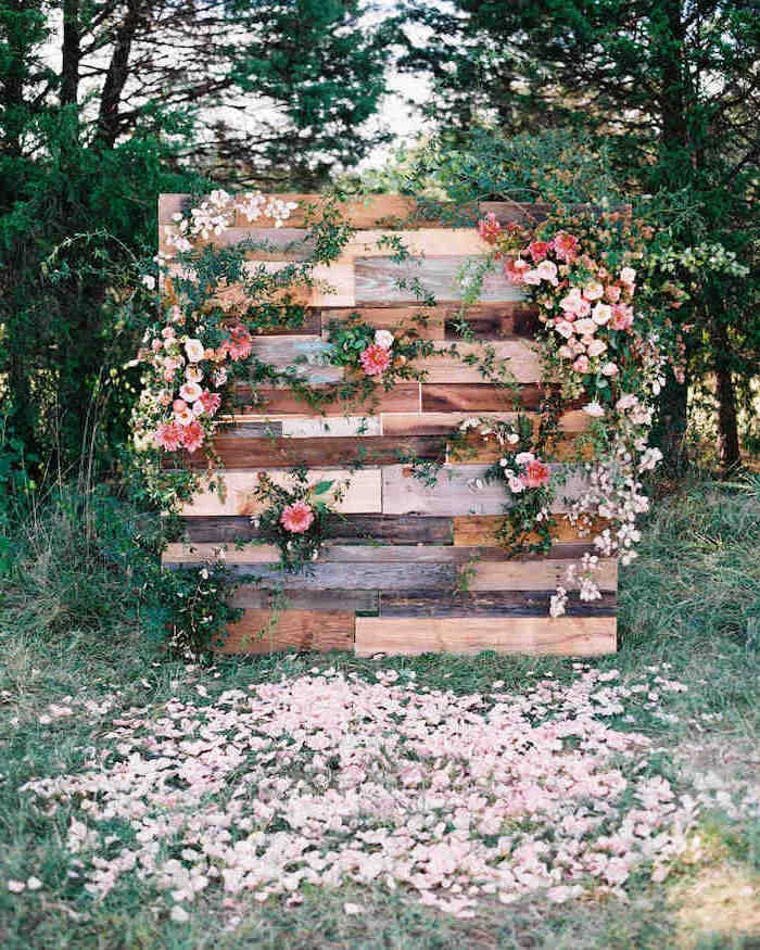 wooden backdrop with pink and white flowers hanging, rose petals in the grass, trees in the background, rustic wedding ideas
