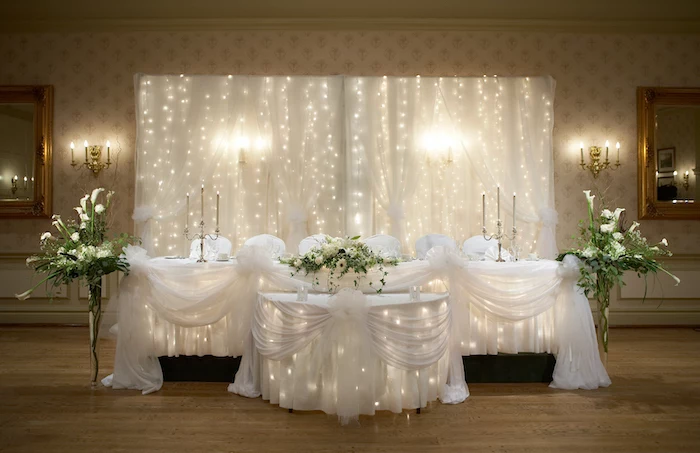 white tulle with fairy lights backdrop, white and green flower arrangements, candelabrum with candles, rustic wedding centrepieces