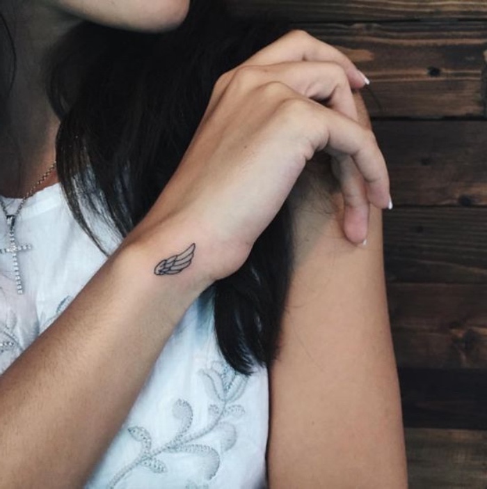 angel wing wrist tattoo, small tattoo ideas, girl wearing a white top, silver cross necklace